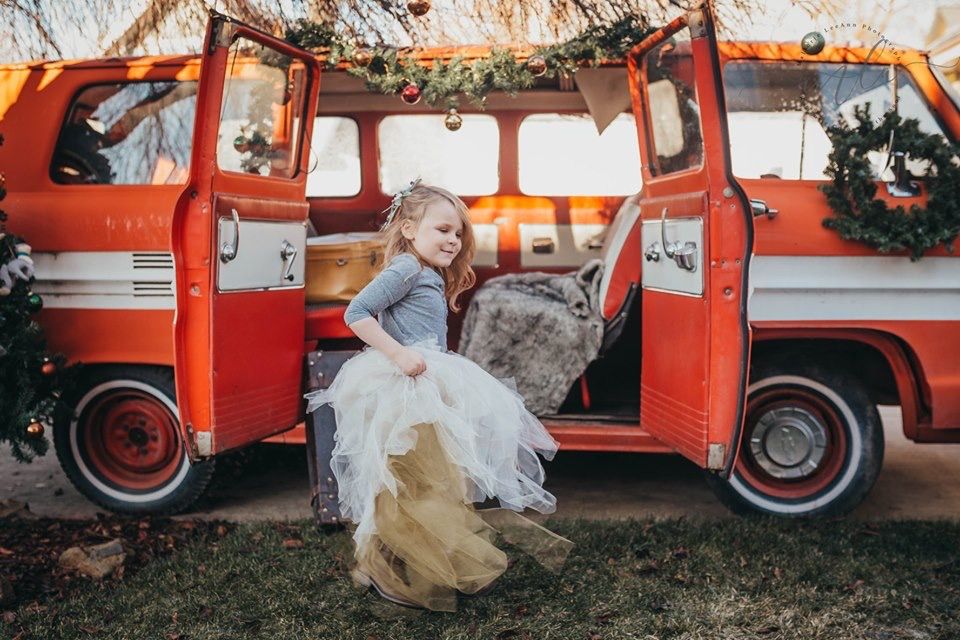 Young girl in white tutu dress plays beside a vintage red Volkswagen van decorated with Christmas wreaths.