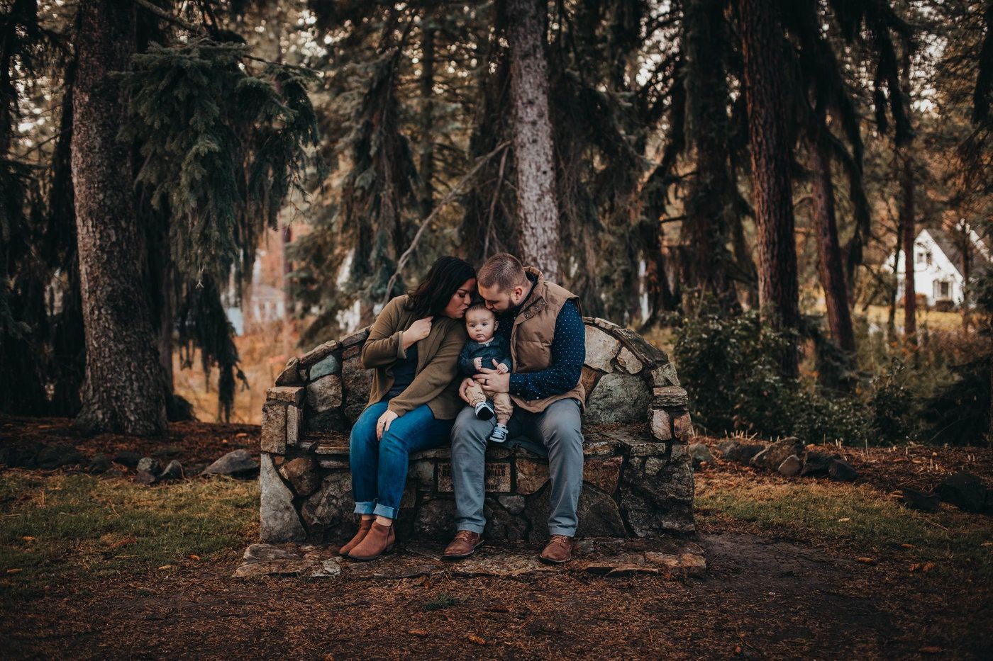 Couple with baby sitting on a rustic stone bench surrounded by tall pine trees in an autumn forest setting.