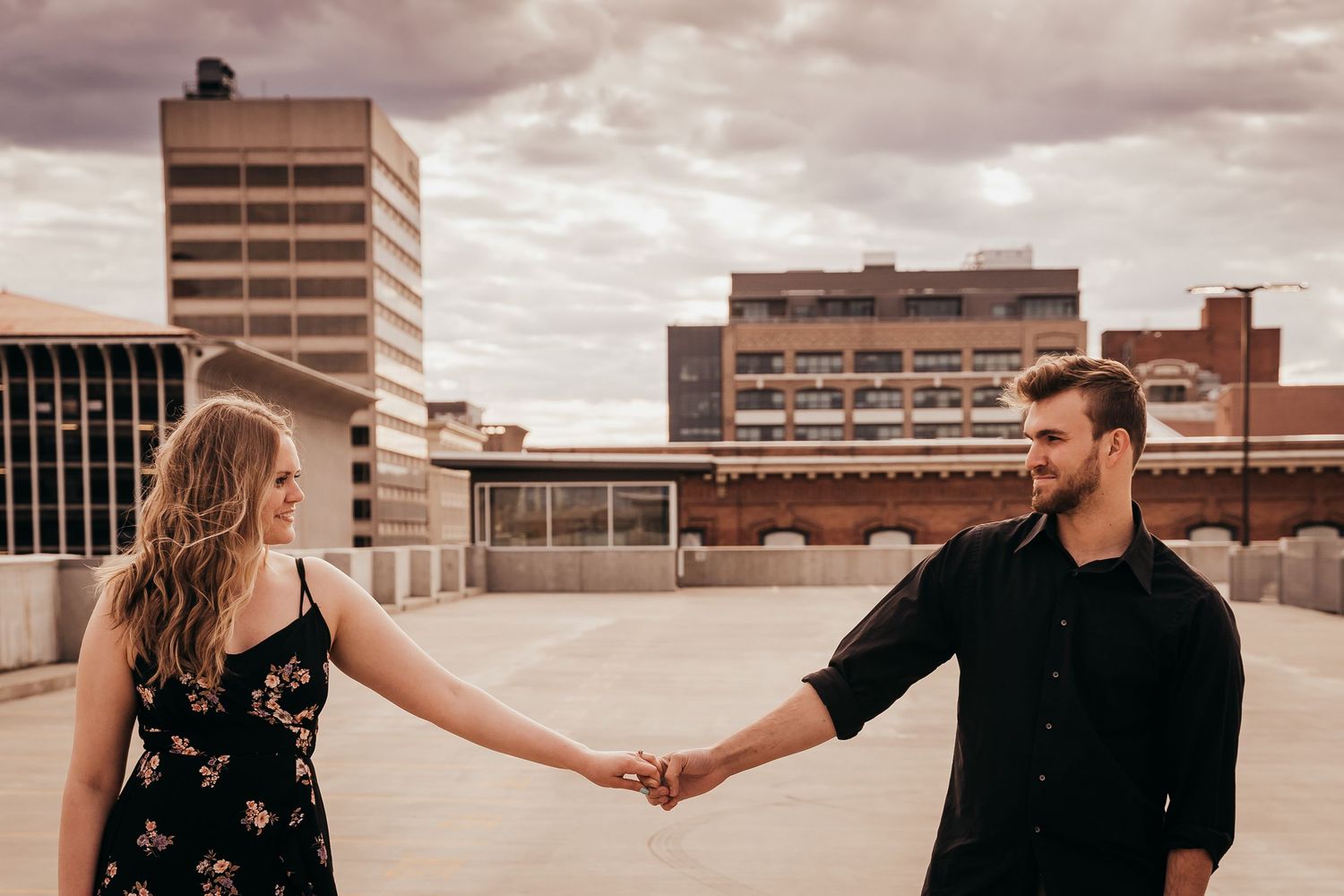 Couple holding hands facing each other on rooftop with cloudy urban skyline behind them.