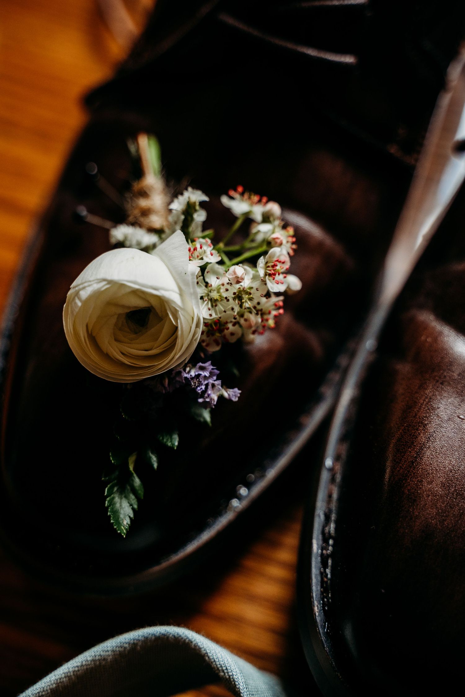 White ranunculus boutonniere resting inside a dark leather dress shoe on a wooden floor.