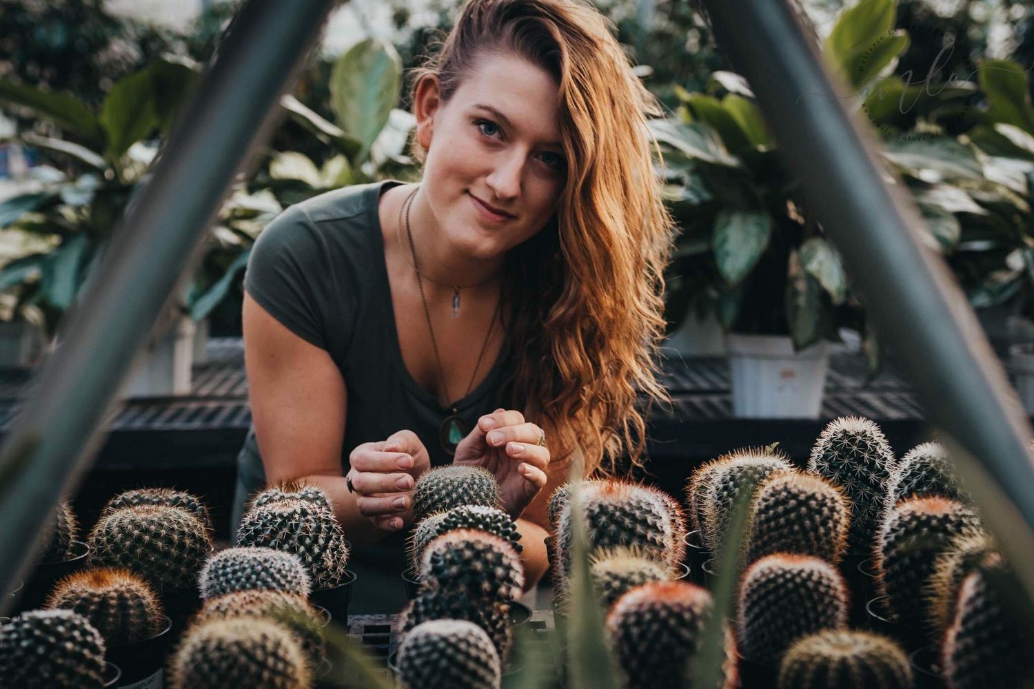 Smiling woman leaning over a display of cacti in a lush greenhouse setting.