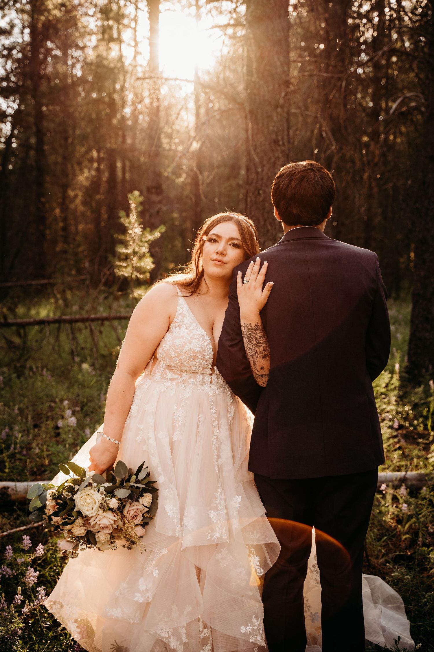 Bride in lace gown holds bouquet leaning on groom in dark suit during golden hour forest wedding portrait.