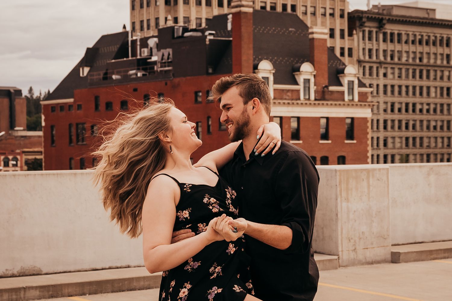 Couple dancing and holding hands on rooftop with urban brick buildings behind them.