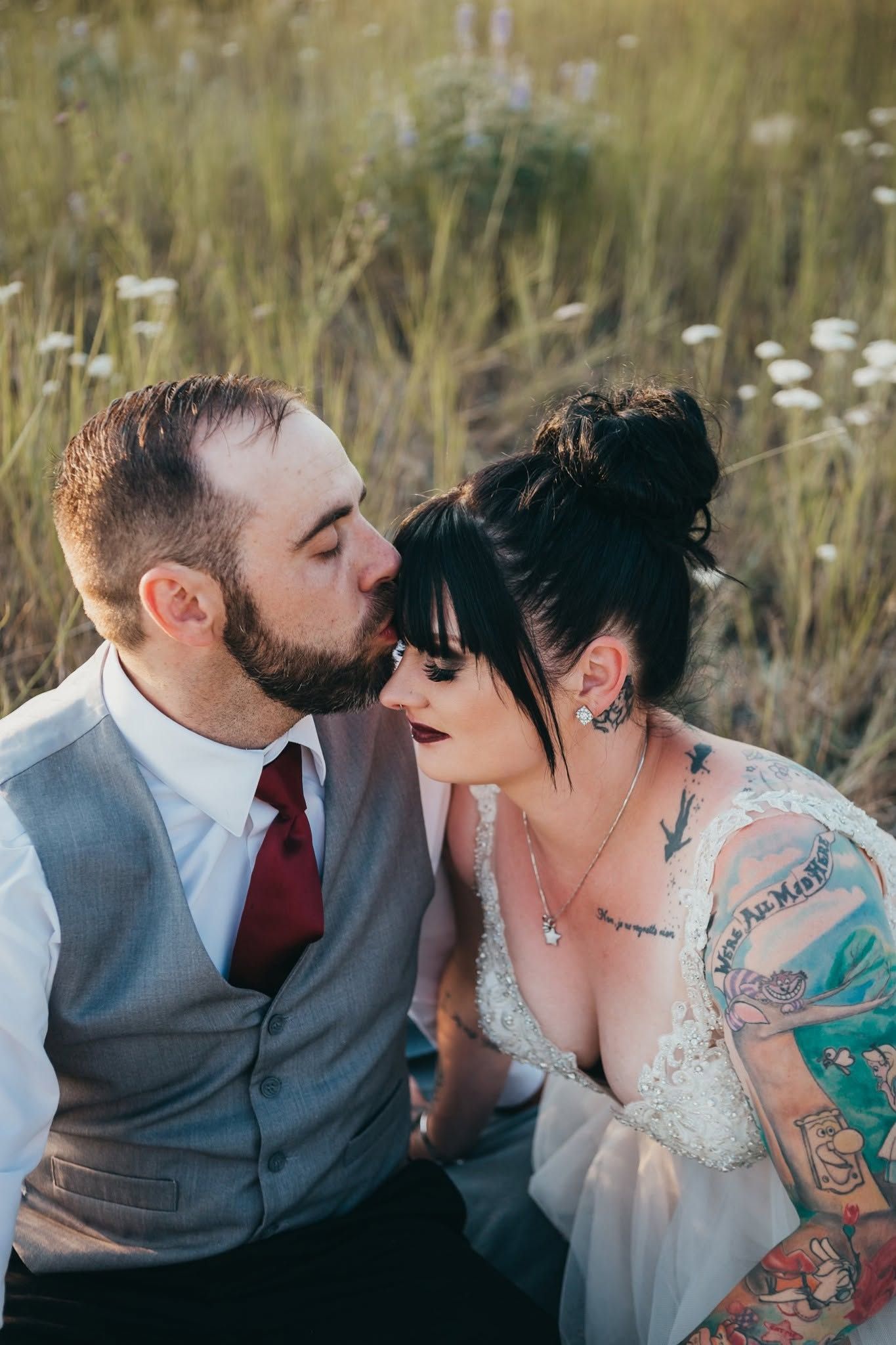Tattooed bride and bearded groom share a tender kiss in a wildflower meadow at sunset.