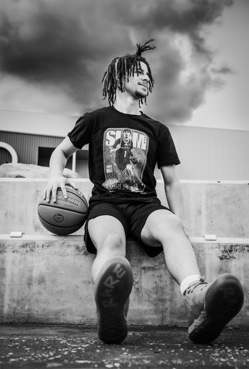 Young man with dreadlocks sitting on concrete ledge holding basketball under dramatic cloudy sky.