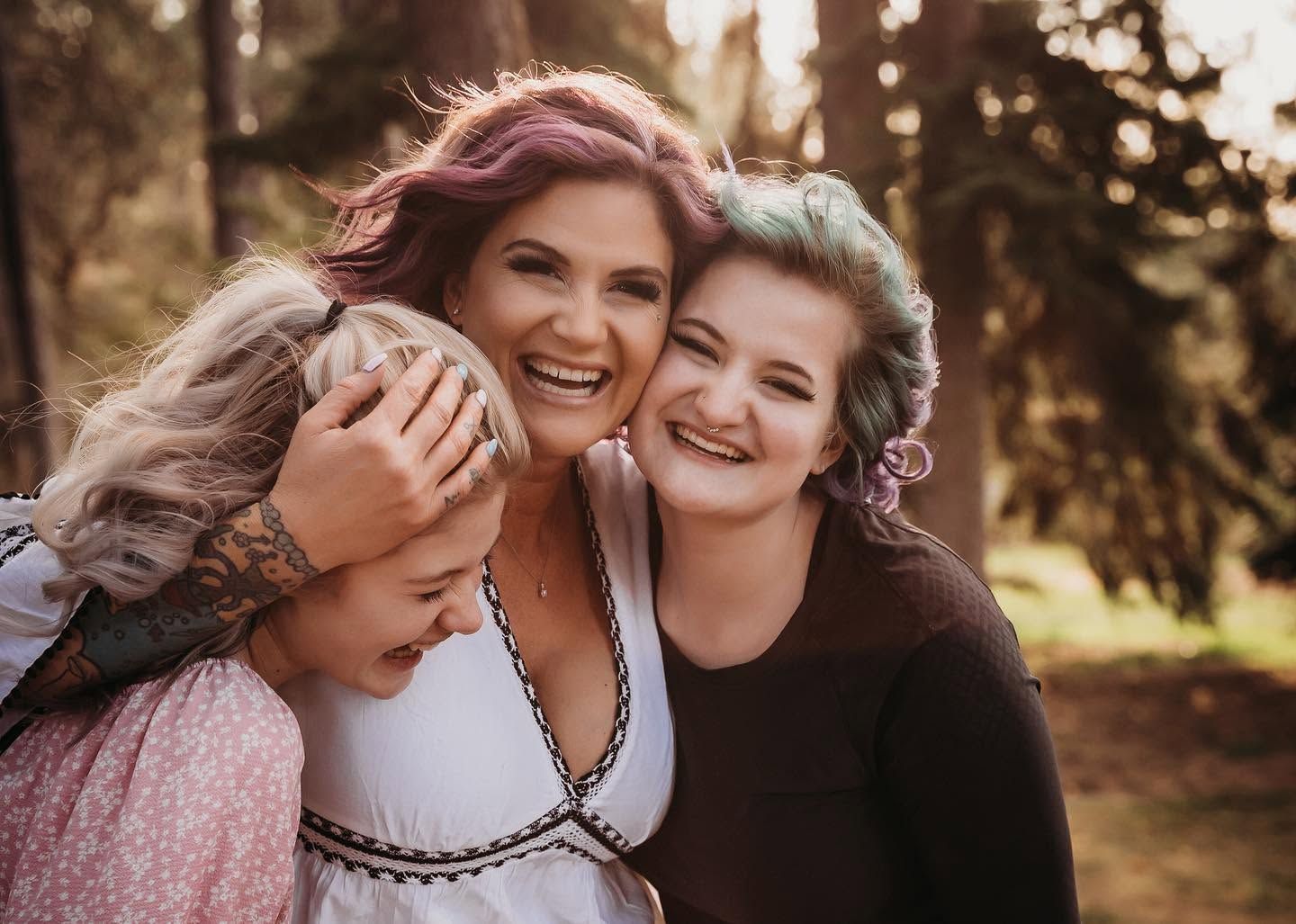 Three women with colorful hair laughing and hugging outdoors in forest.