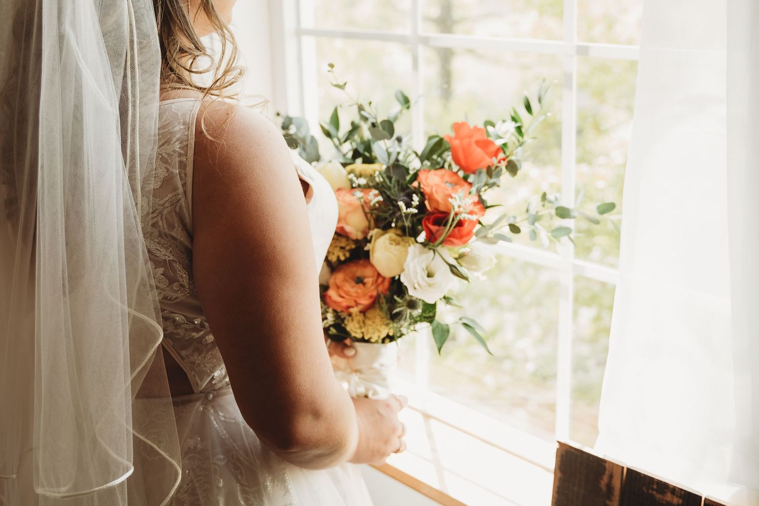 Bride in lace wedding gown holding colorful orange and white bouquet by a sunlit window.