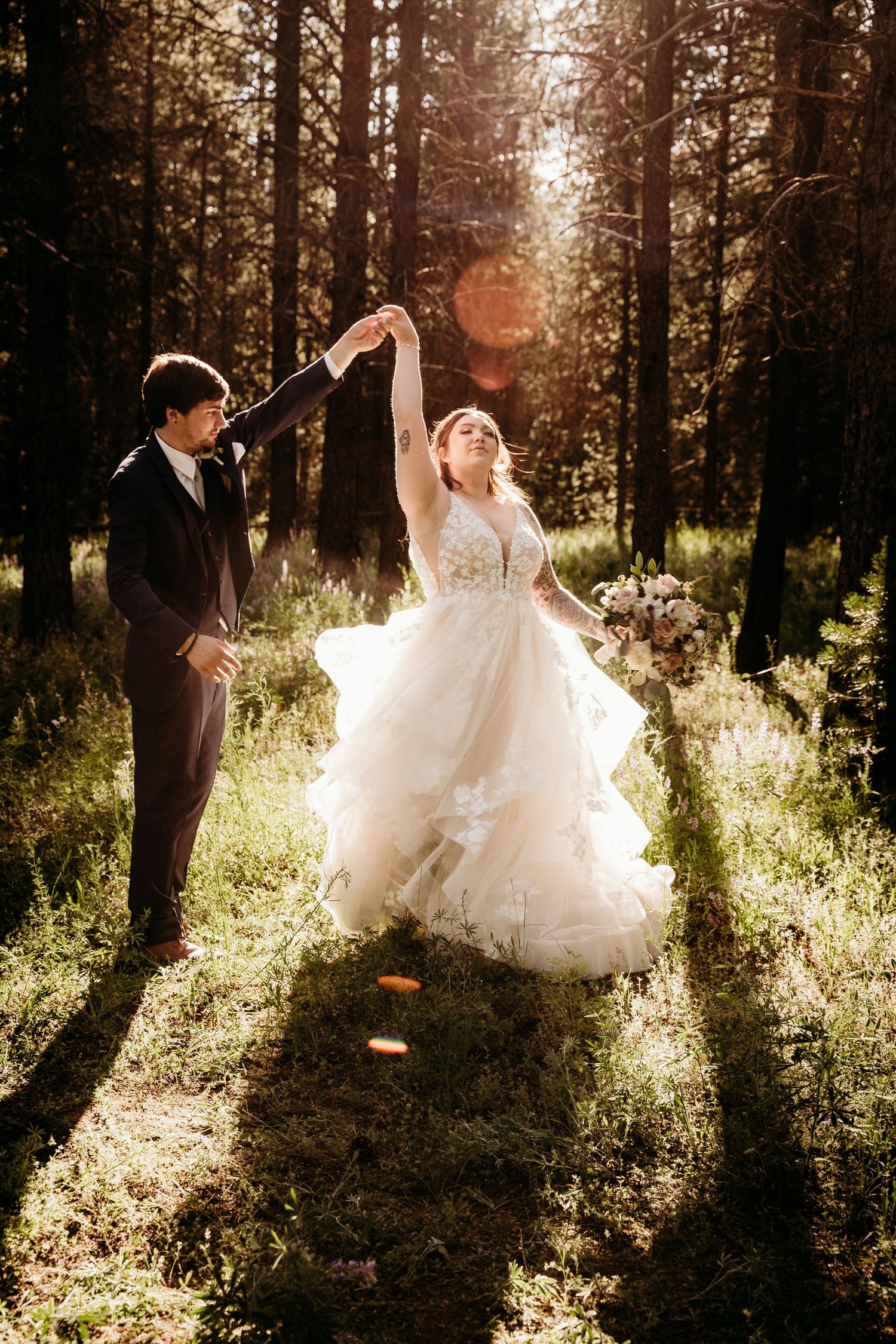 Groom twirls bride in voluminous white ruffled gown in dramatic sunlit forest with beautiful lens flare.