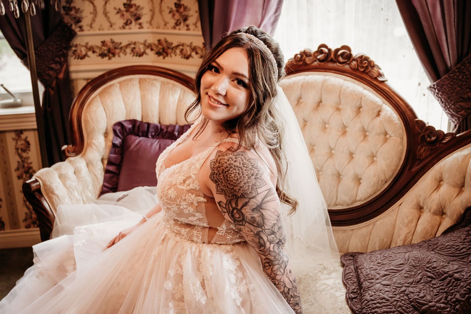 Smiling tattooed bride in a blush lace wedding gown with veil seated on a tufted vintage sofa.