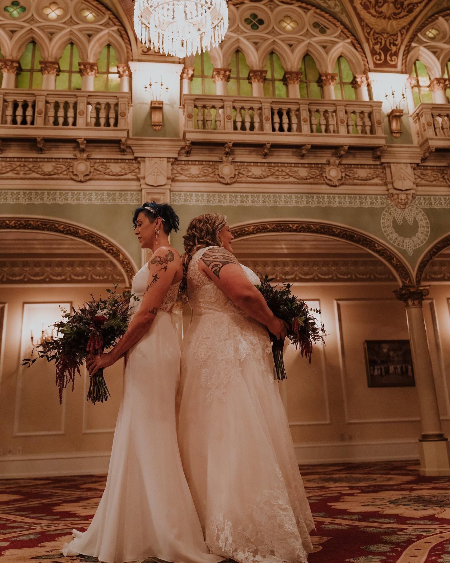 Two brides in white lace gowns stand back-to-back holding bouquets inside an ornate grand ballroom venue.