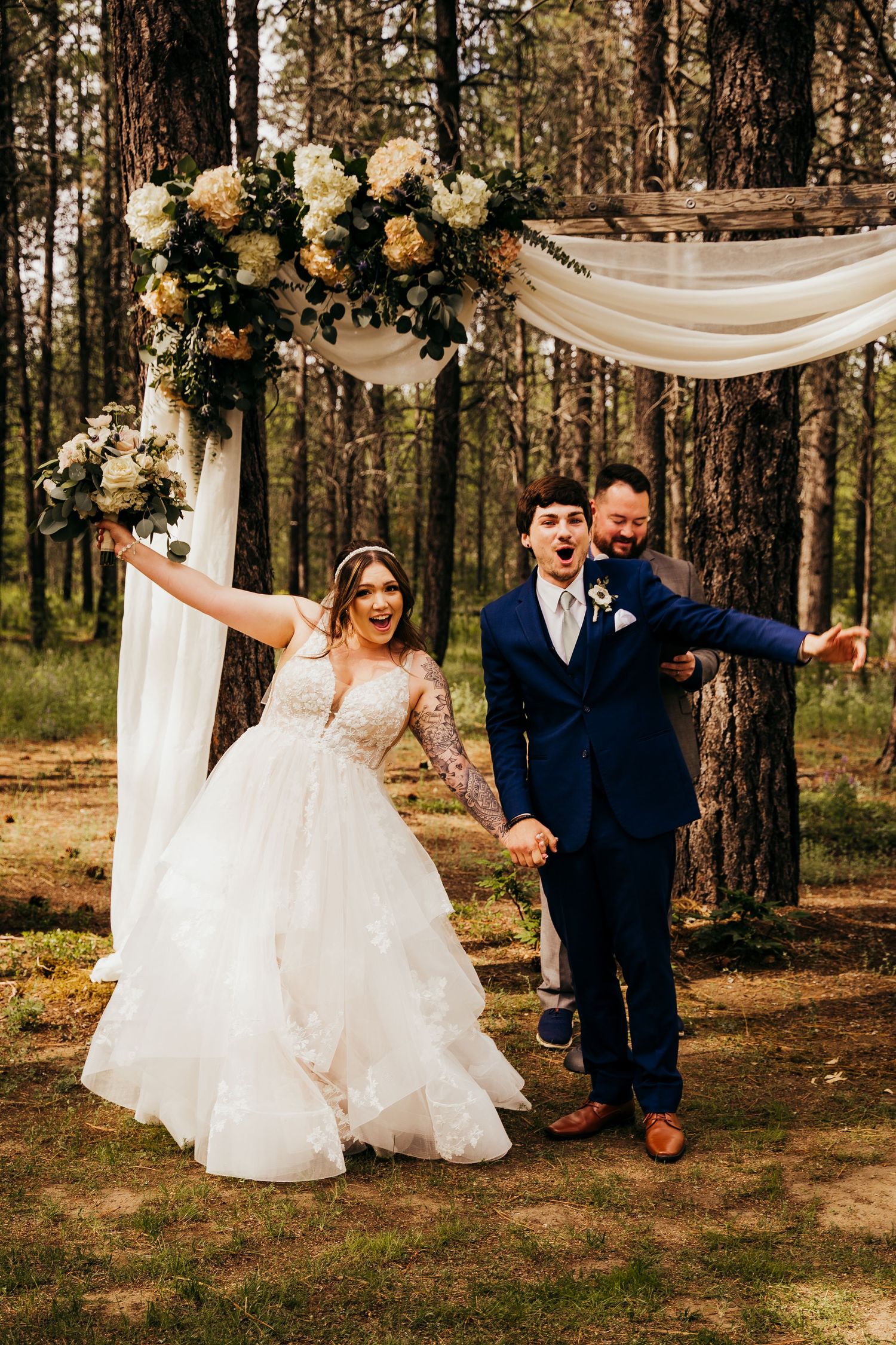 Joyful bride and groom celebrating after ceremony beneath floral arch in pine forest.