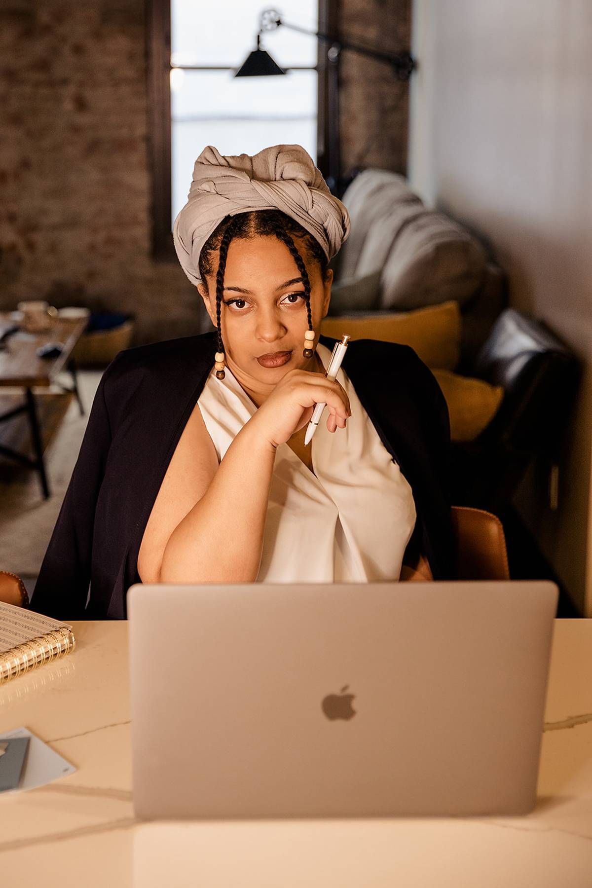 Black woman wearing a white head wrap sitting at a desk with an Apple MacBook laptop in a loft office.