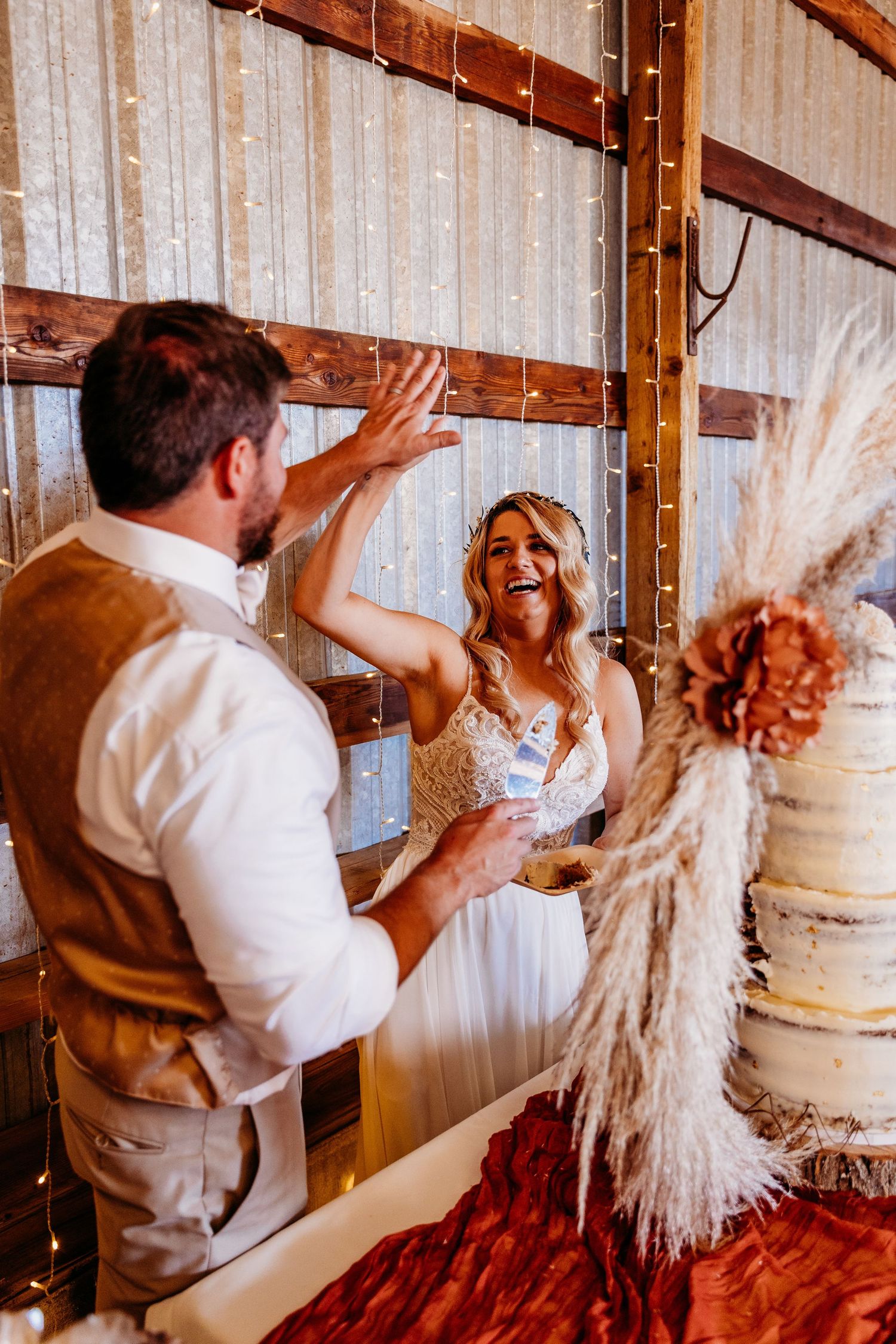 Bride and groom high-five joyfully during wedding cake cutting in rustic barn.