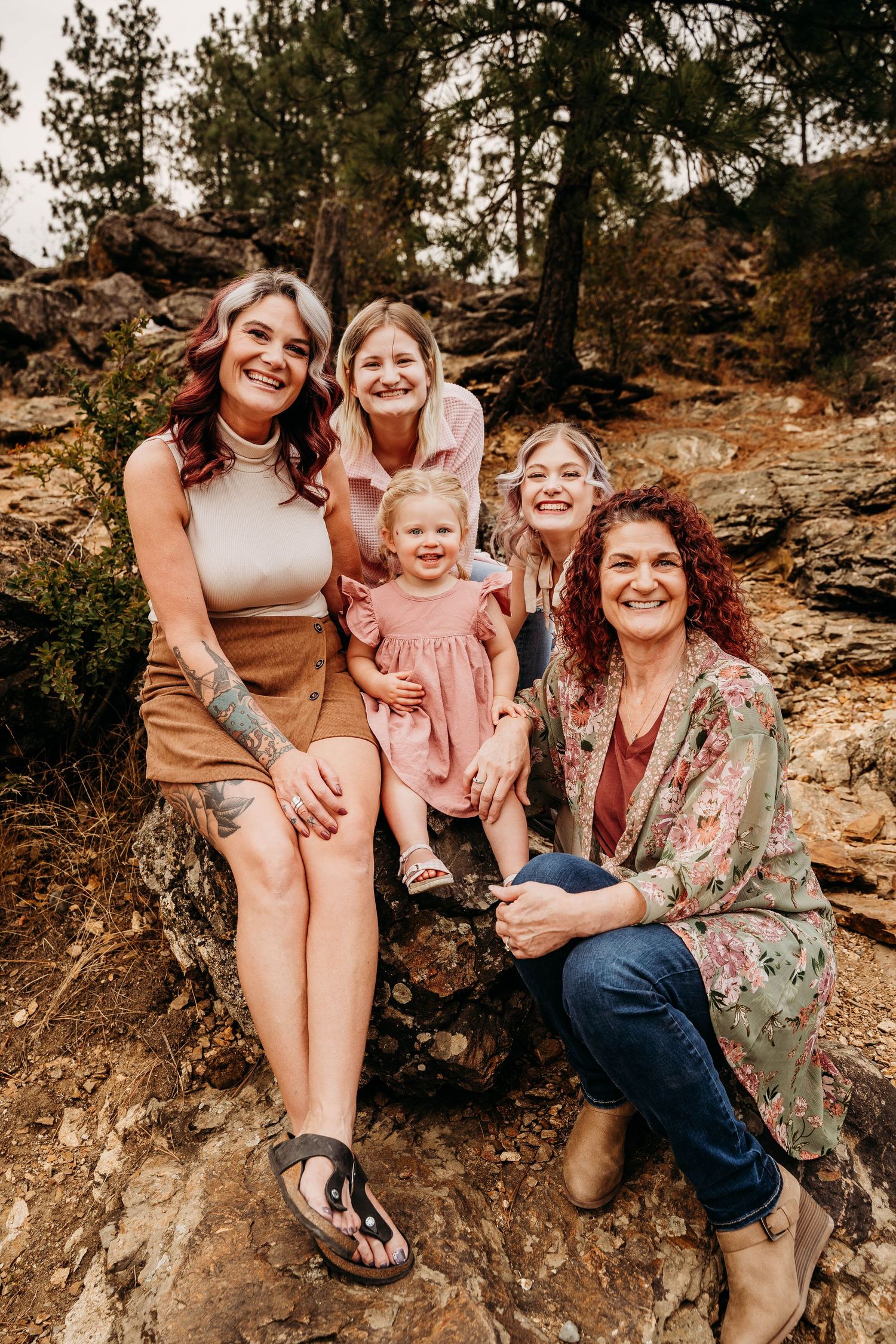Four women and a toddler pose together on rocks among pine trees, smiling for a candid family portrait.