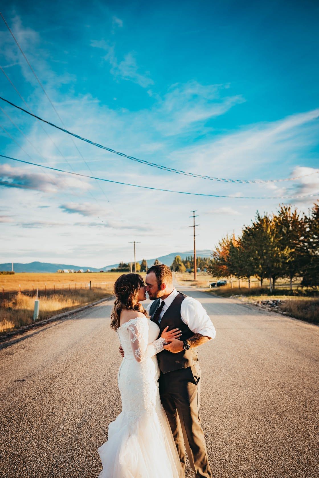 Bride and groom kiss on an open rural road under a bright blue sky on wedding day.
