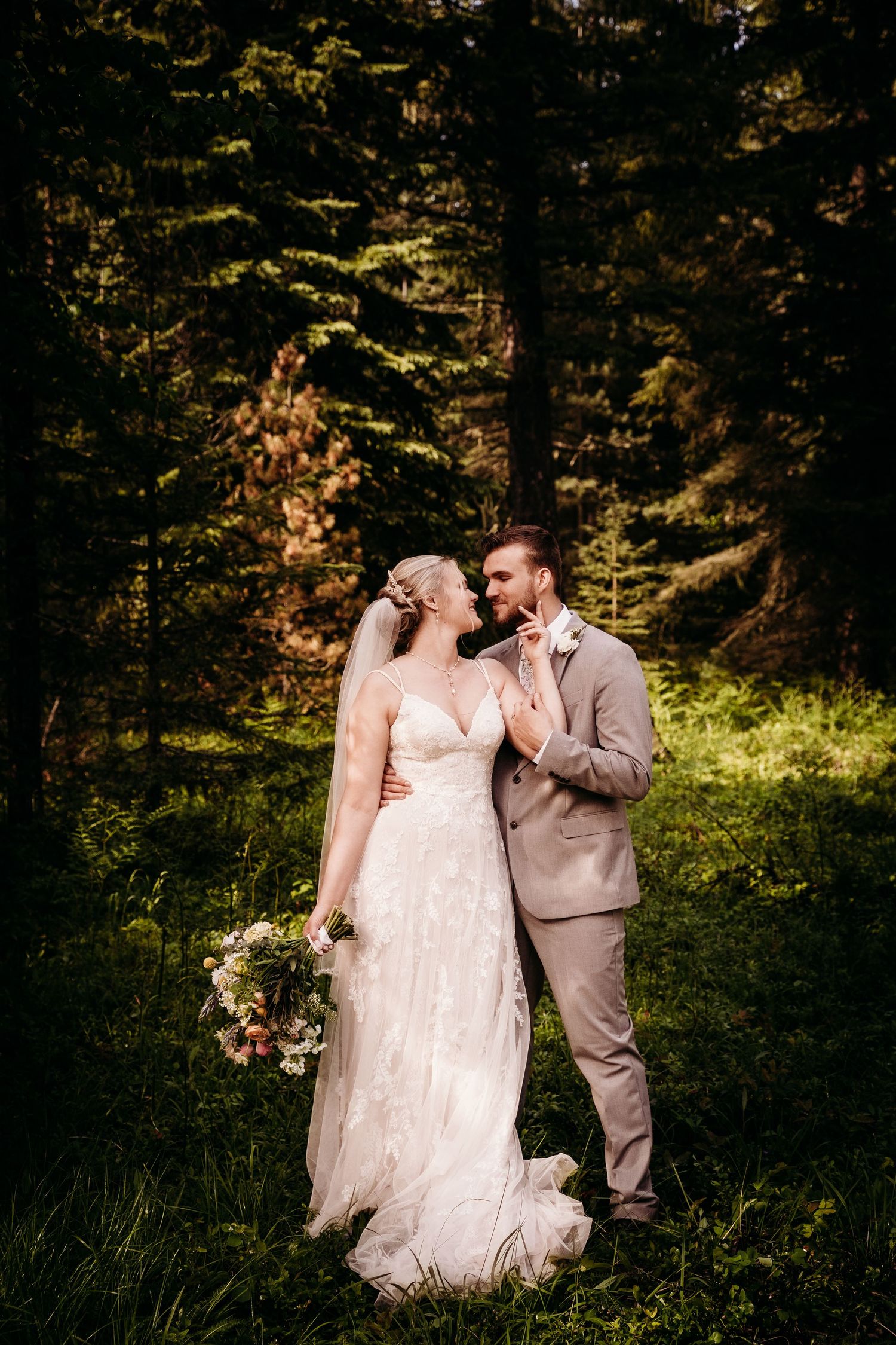 Bride and groom standing close in a dark forest setting, bride holding a floral bouquet.