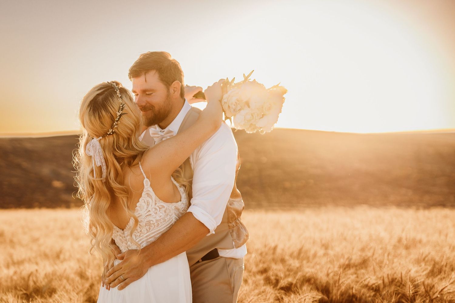 Couple shares a romantic kiss in a golden wheat field at sunset on their wedding day.
