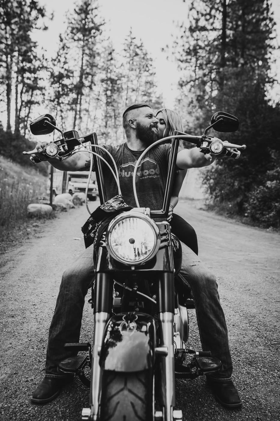 Couple poses on a classic motorcycle on a tree-lined road in a black and white photo.