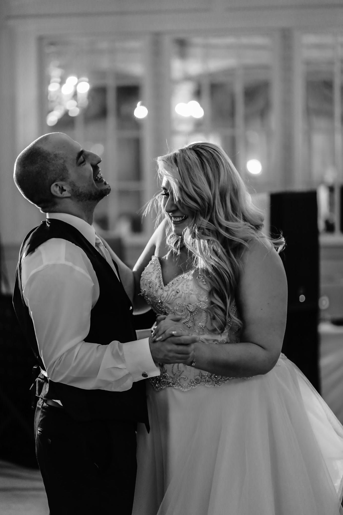 Bride and groom share joyful first dance in elegant ballroom in black and white photo.