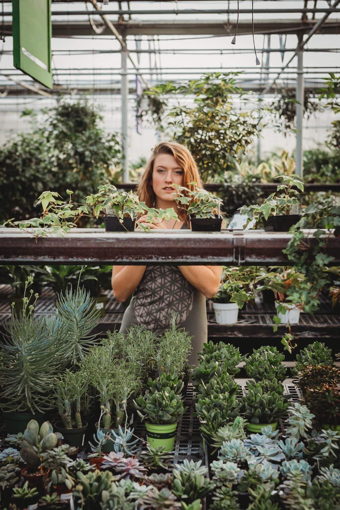 Young woman with auburn hair stands among rows of potted plants and succulents in greenhouse.