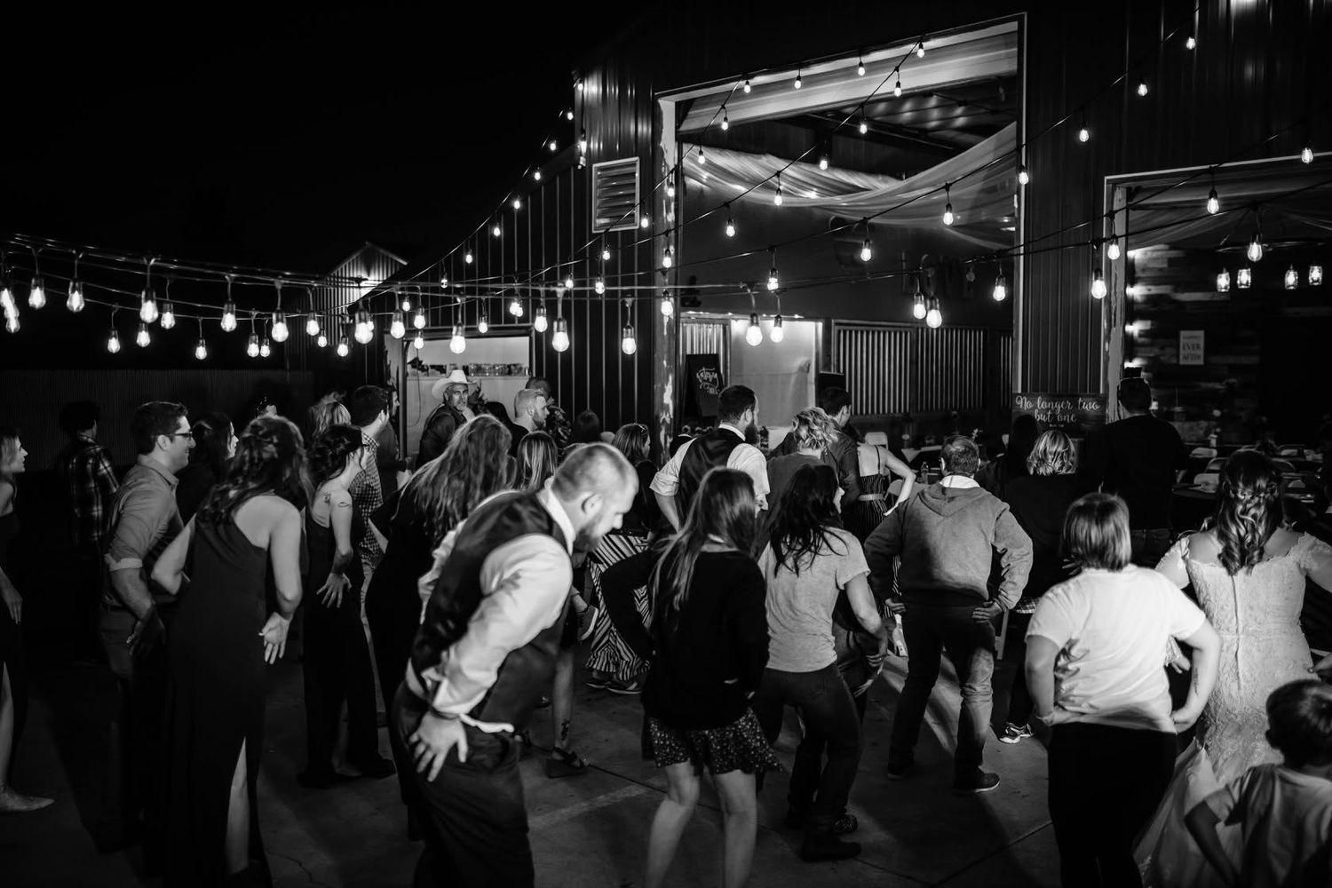 Guests dance under string lights at an outdoor wedding reception in black and white photo.