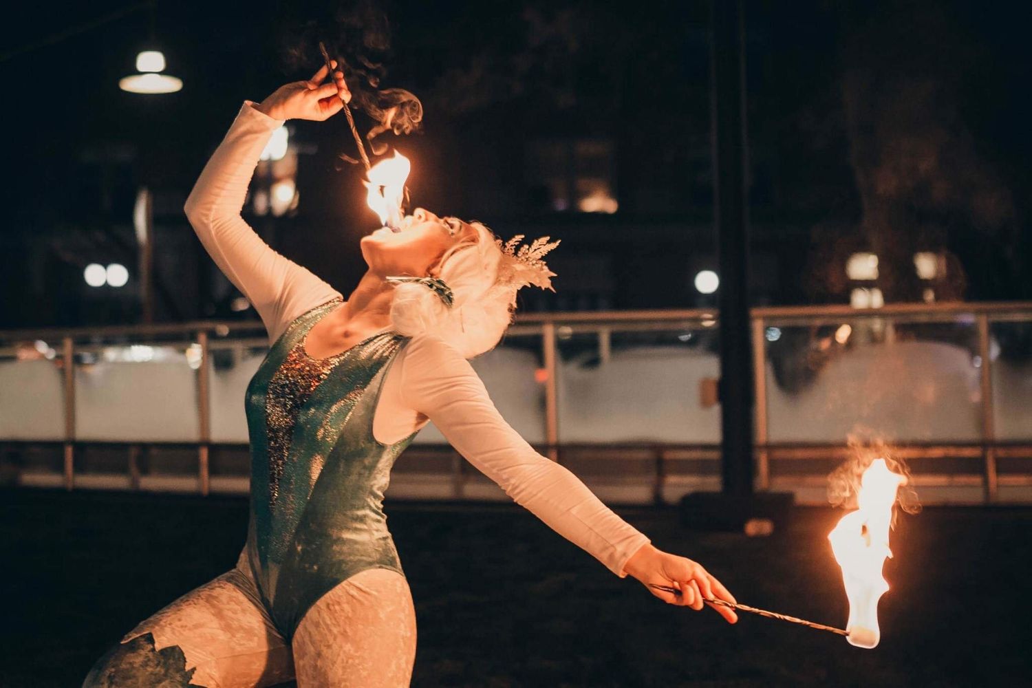 Female fire dancer in green costume bending back while performing with flames at night.