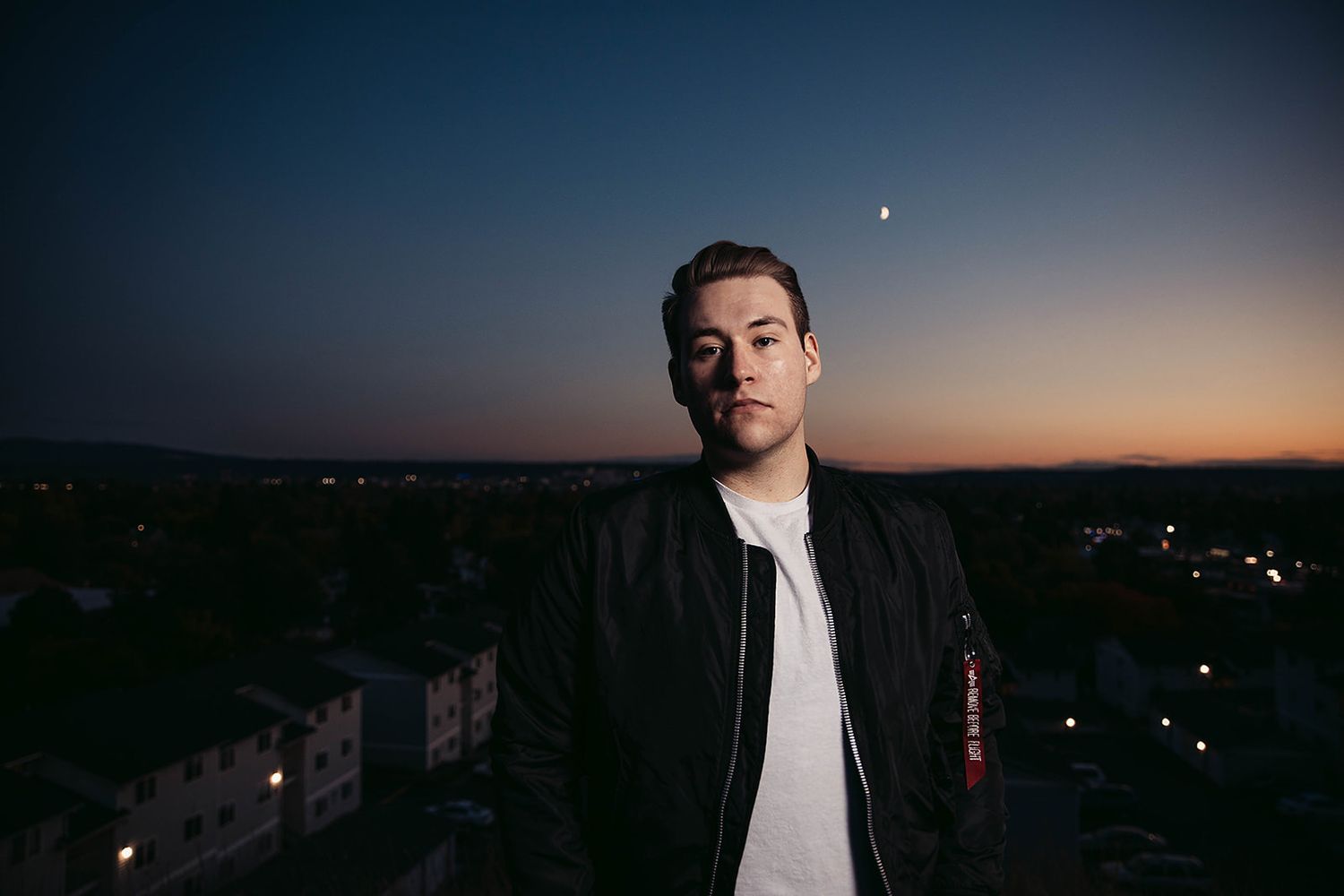Man in black jacket standing on rooftop at dusk with city lights below.