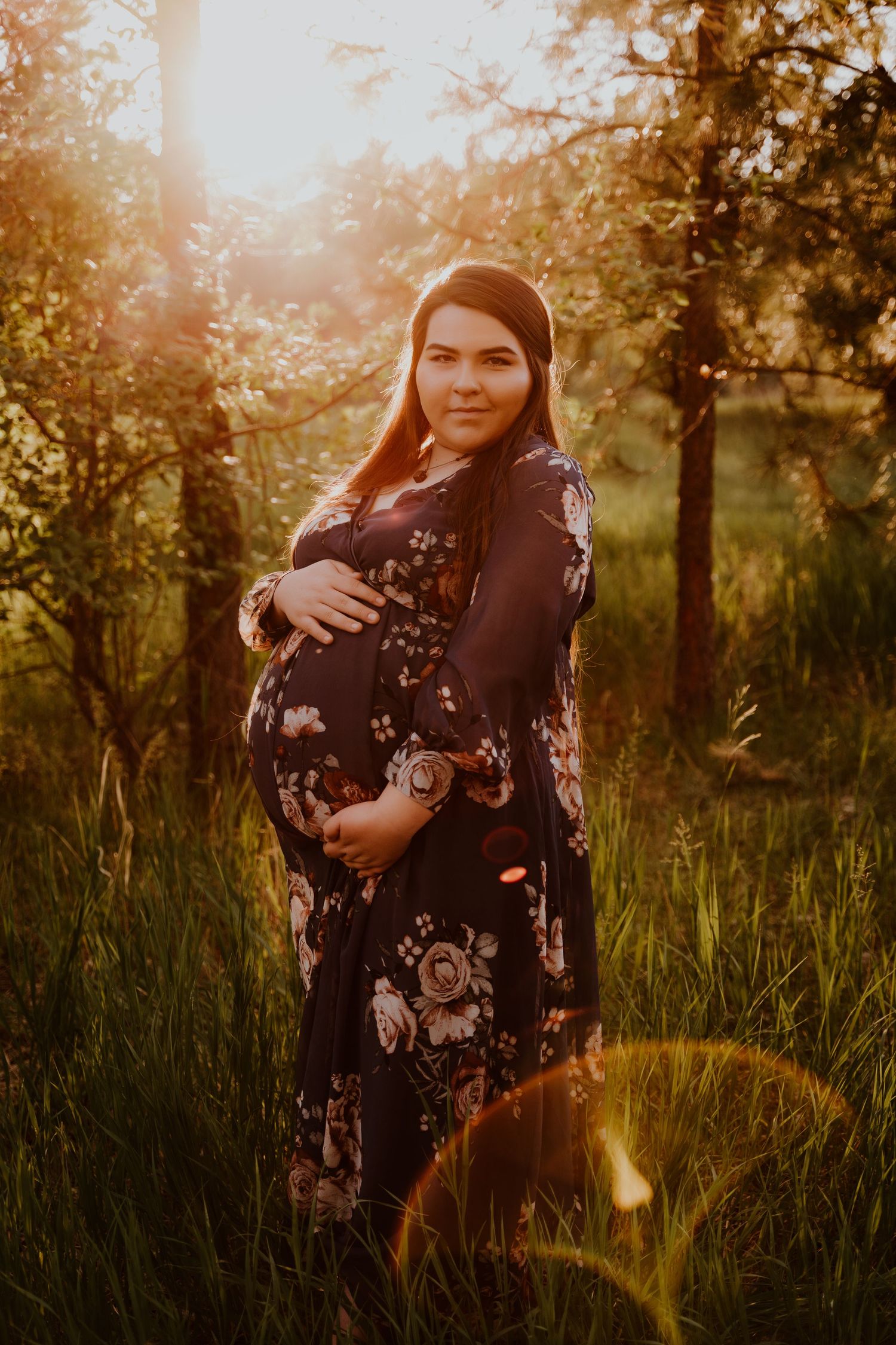 Pregnant woman in floral dress cradling baby bump during golden hour sunset in forest.