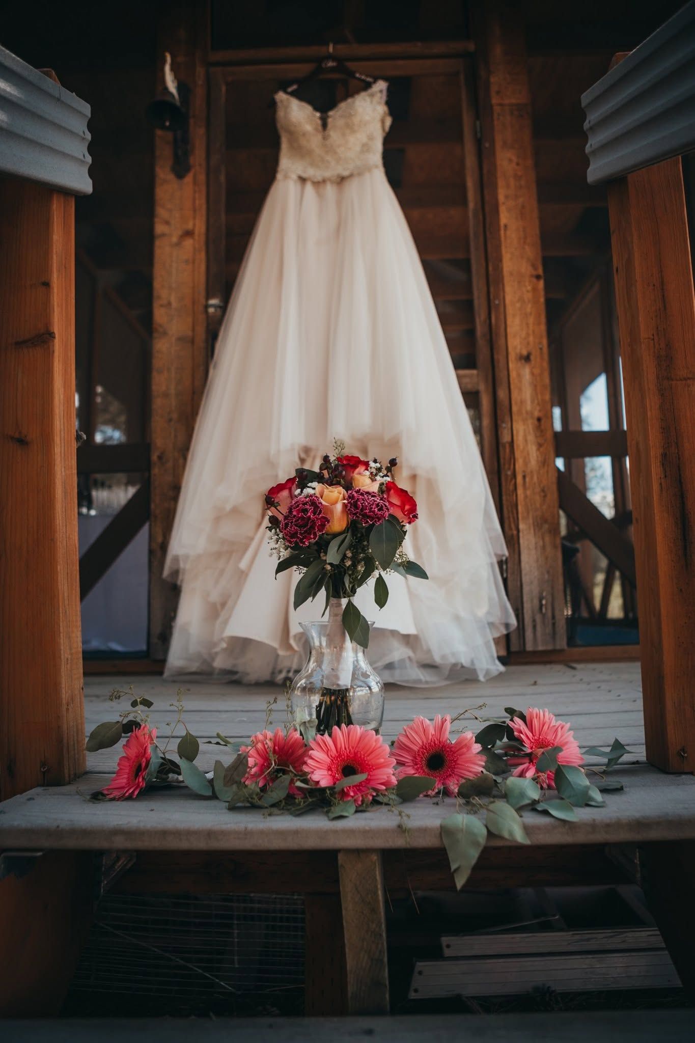 Elegant white ball gown wedding dress hanging with colorful bridal bouquet on rustic porch.