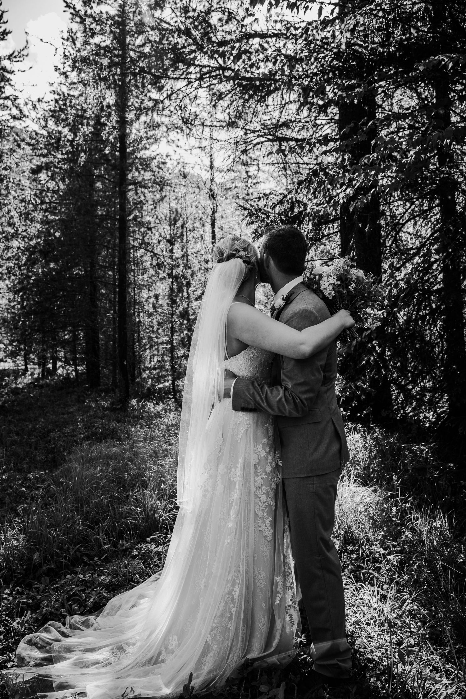 Black and white photo of bride and groom embracing and kissing in a forest setting.