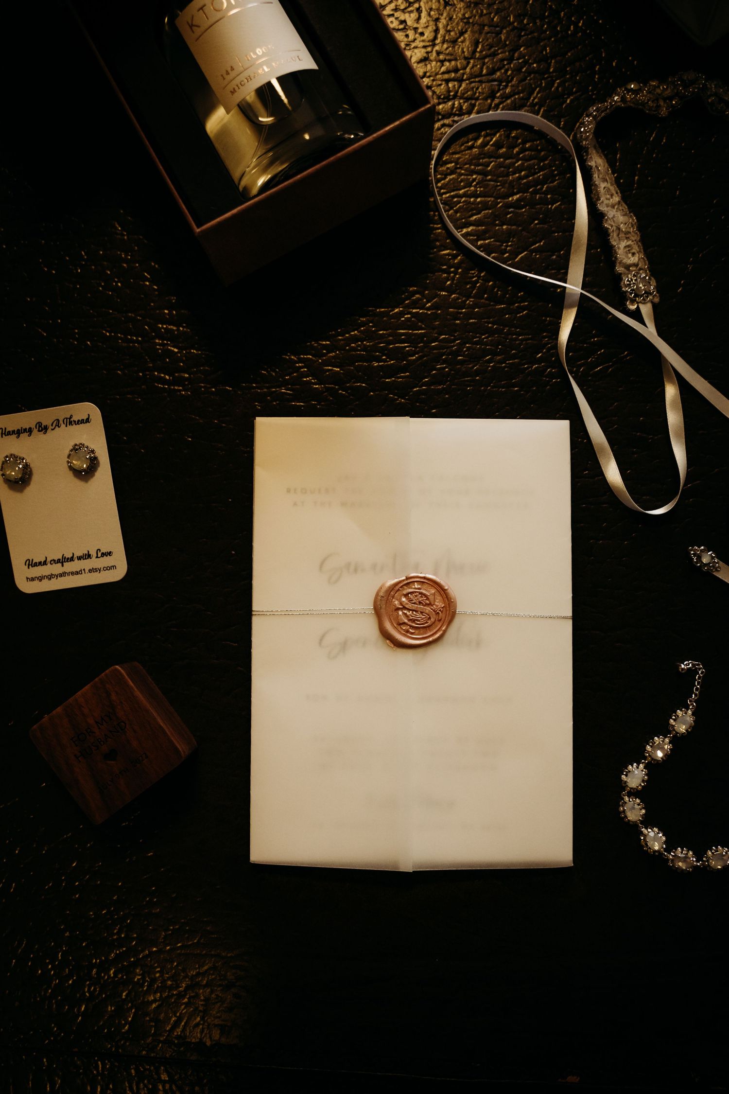 Elegant wax sealed wedding invitation with rose stamp surrounded by jewelry, perfume and ribbon on dark surface.