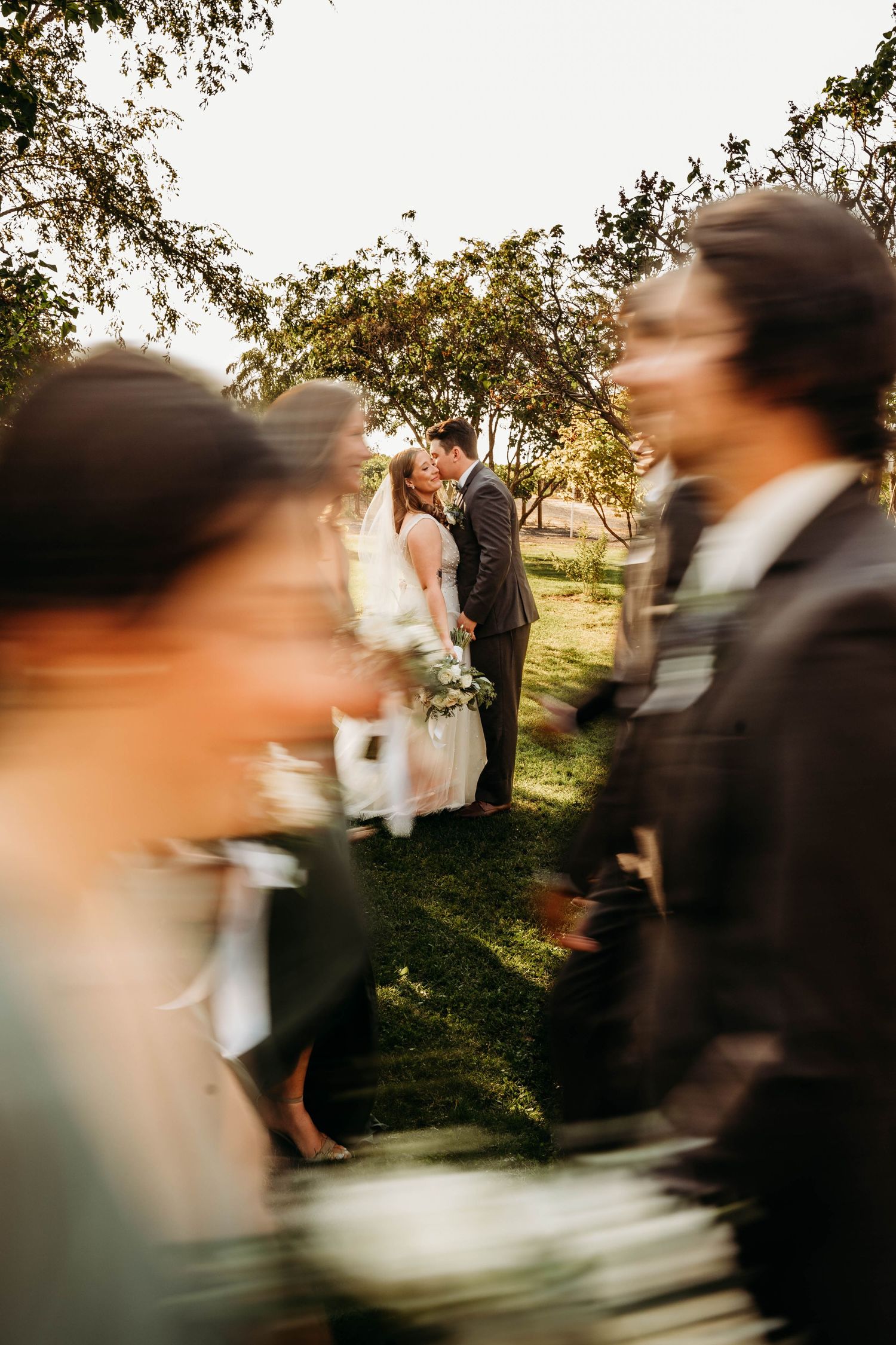 Bride and groom share a kiss in soft golden light while blurred wedding party passes in foreground.
