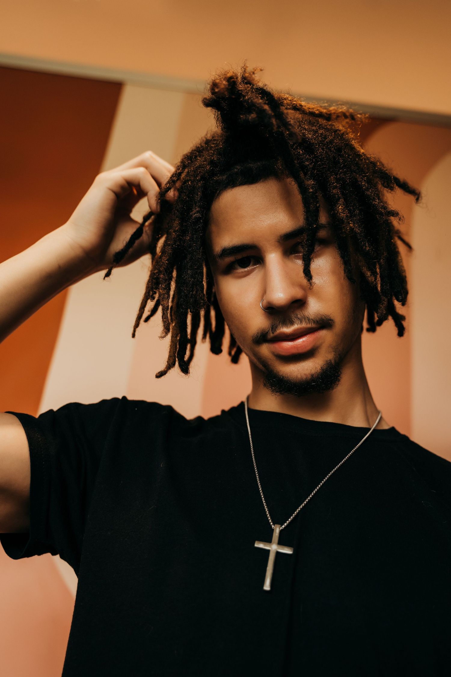Young man with dreadlocks wearing black shirt and cross necklace posing confidently indoors.