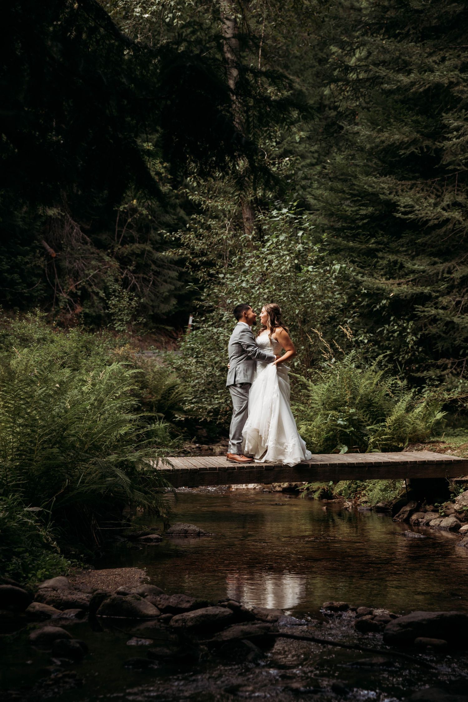 Newlyweds embracing on a wooden bridge over a rocky stream in a dark lush forest setting.