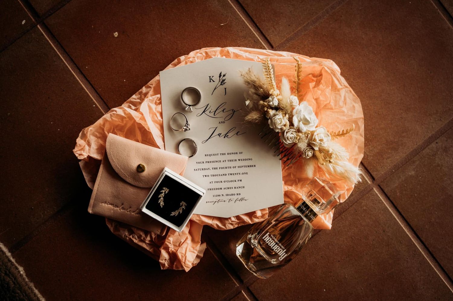 Boho wedding flat lay with invitation, rings, earrings, dried flowers, and perfume on tile.