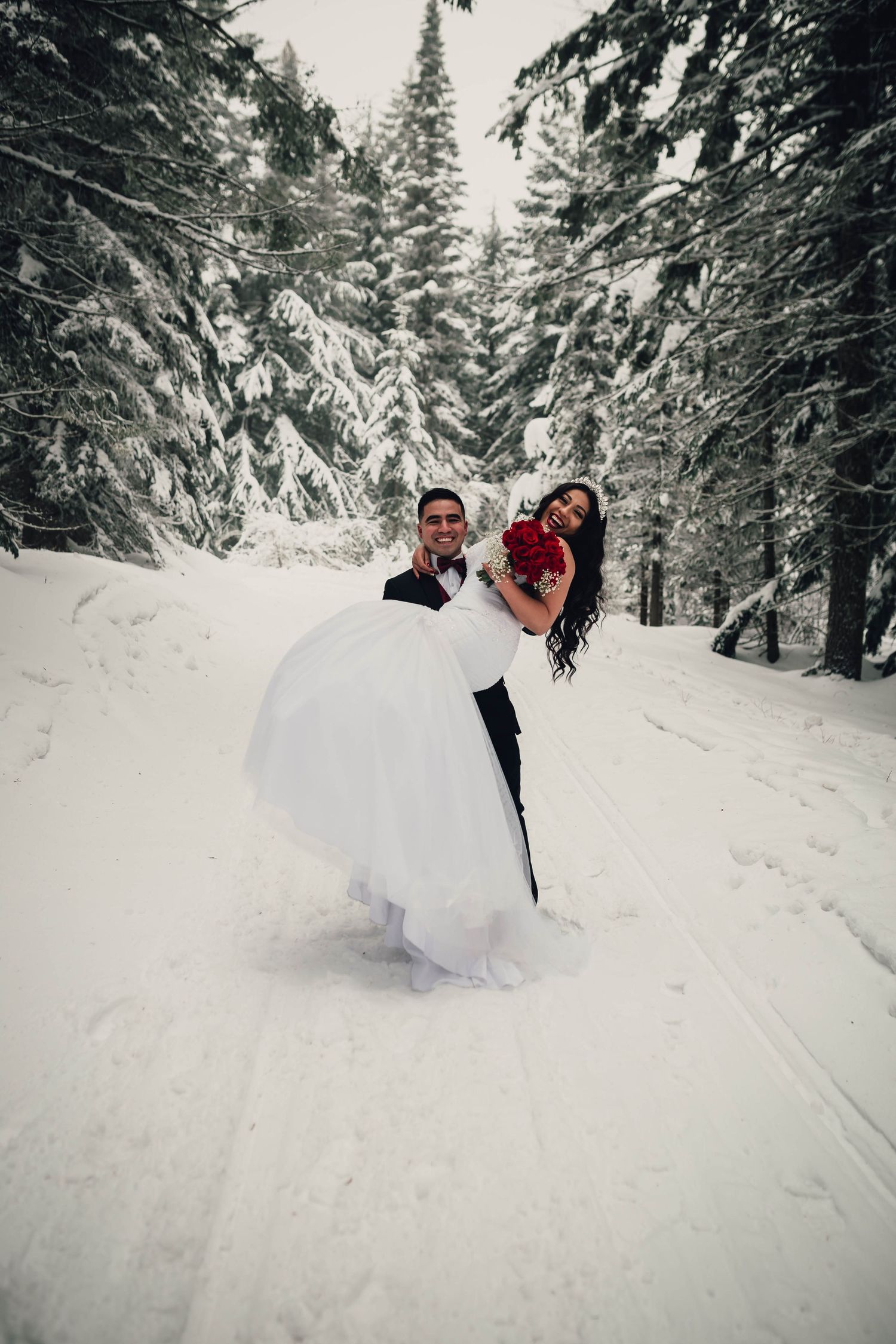 Groom carries laughing bride in white wedding dress through snowy pine forest in winter wonderland setting.