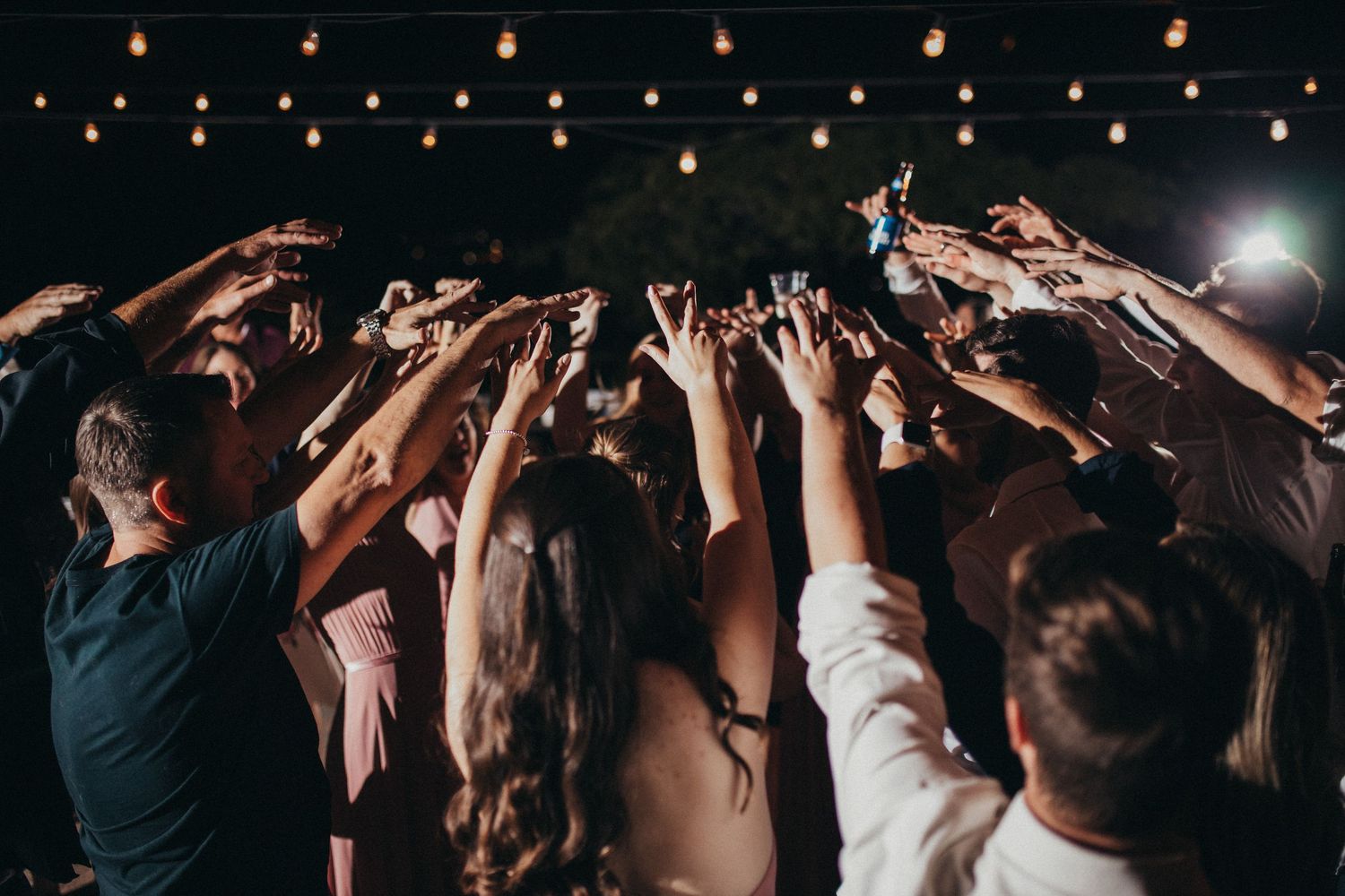 Wedding guests raise their hands and phones in celebration on the dance floor under warm string lights at night.