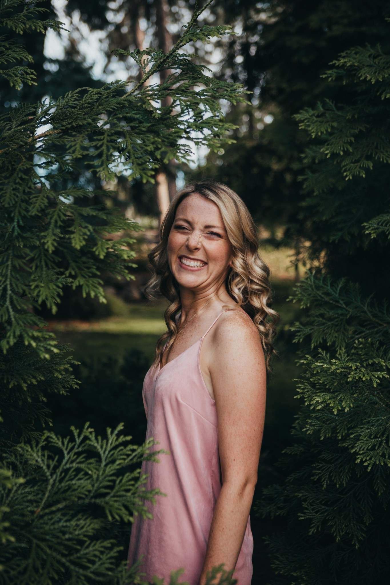 Smiling blonde woman in pink dress standing among lush green cedar trees in a park.