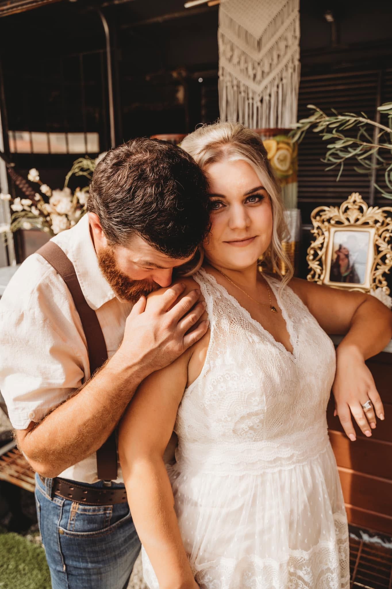 Groom in suspenders nuzzling bride's neck at boho wedding reception with macrame backdrop.