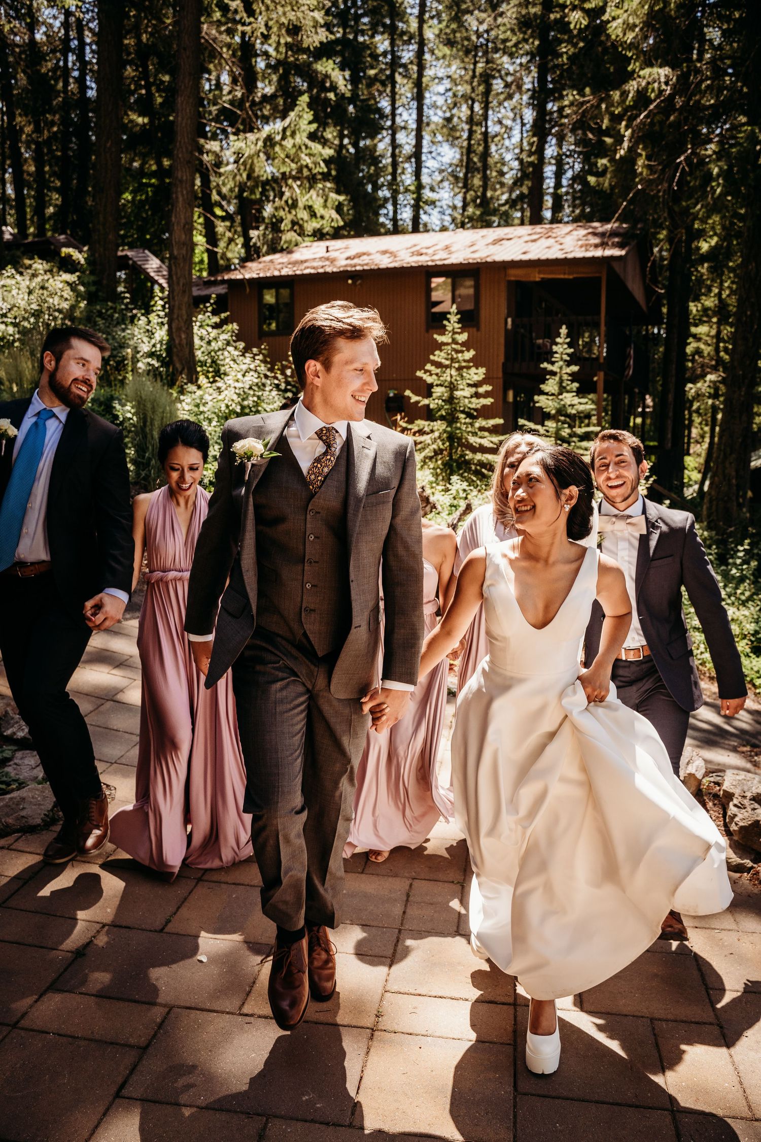 Joyful newlyweds holding hands walking with wedding party through sunlit forest path.