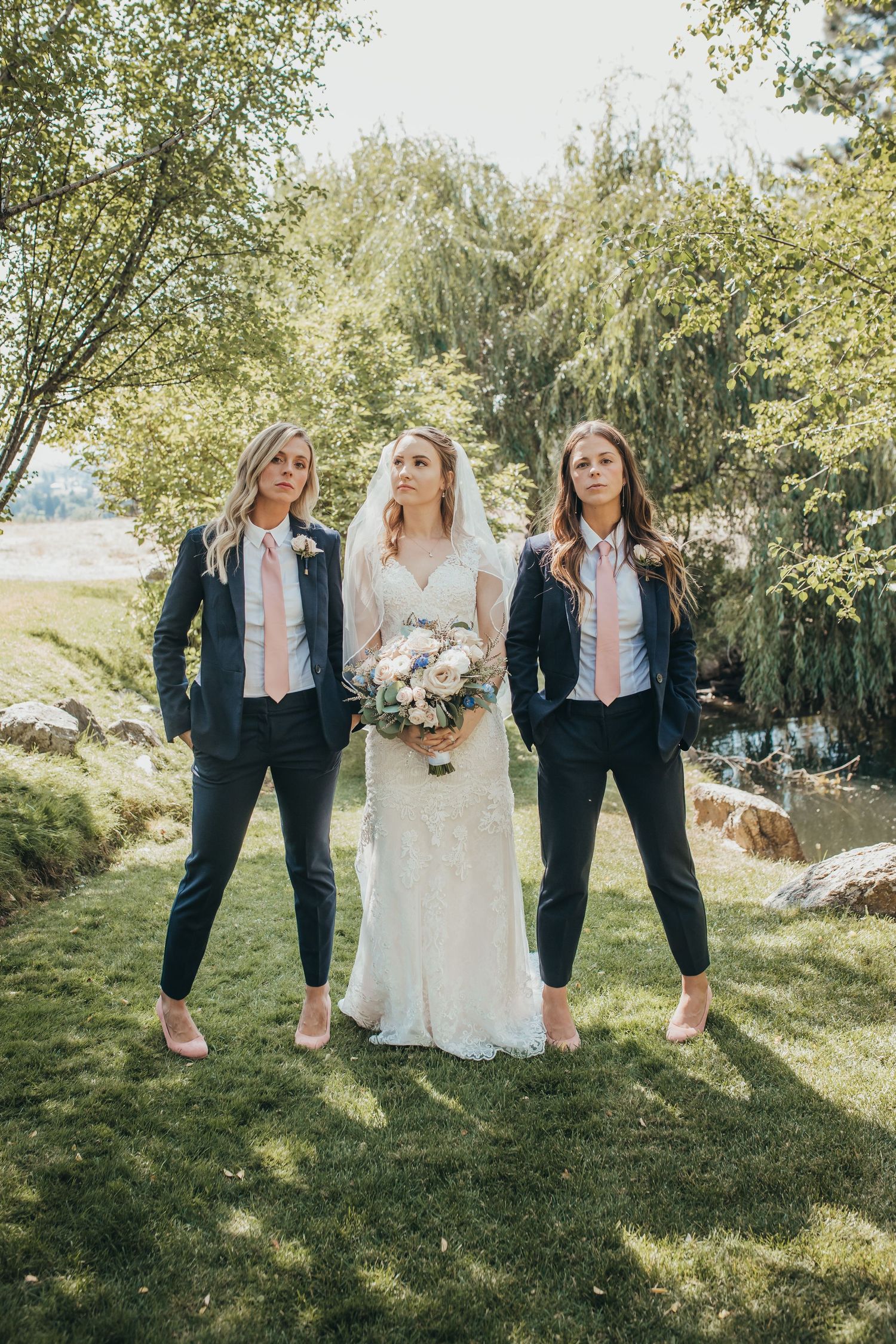 Bride in white gown poses with two bridesmaids wearing navy suits and pink ties outdoors.