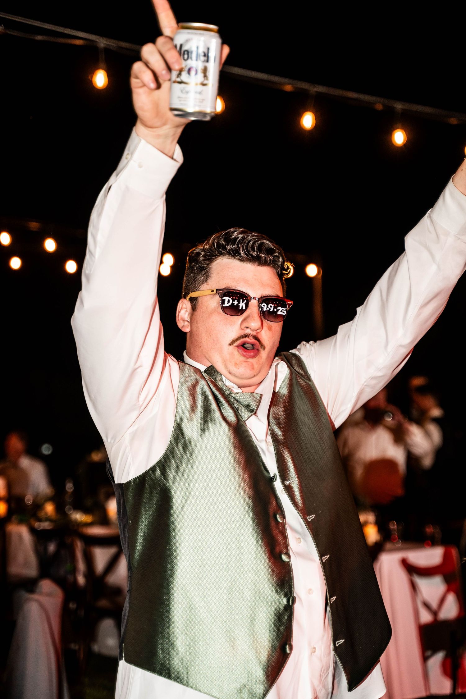 Enthusiastic wedding guest in novelty sunglasses raises a beer can at outdoor reception.