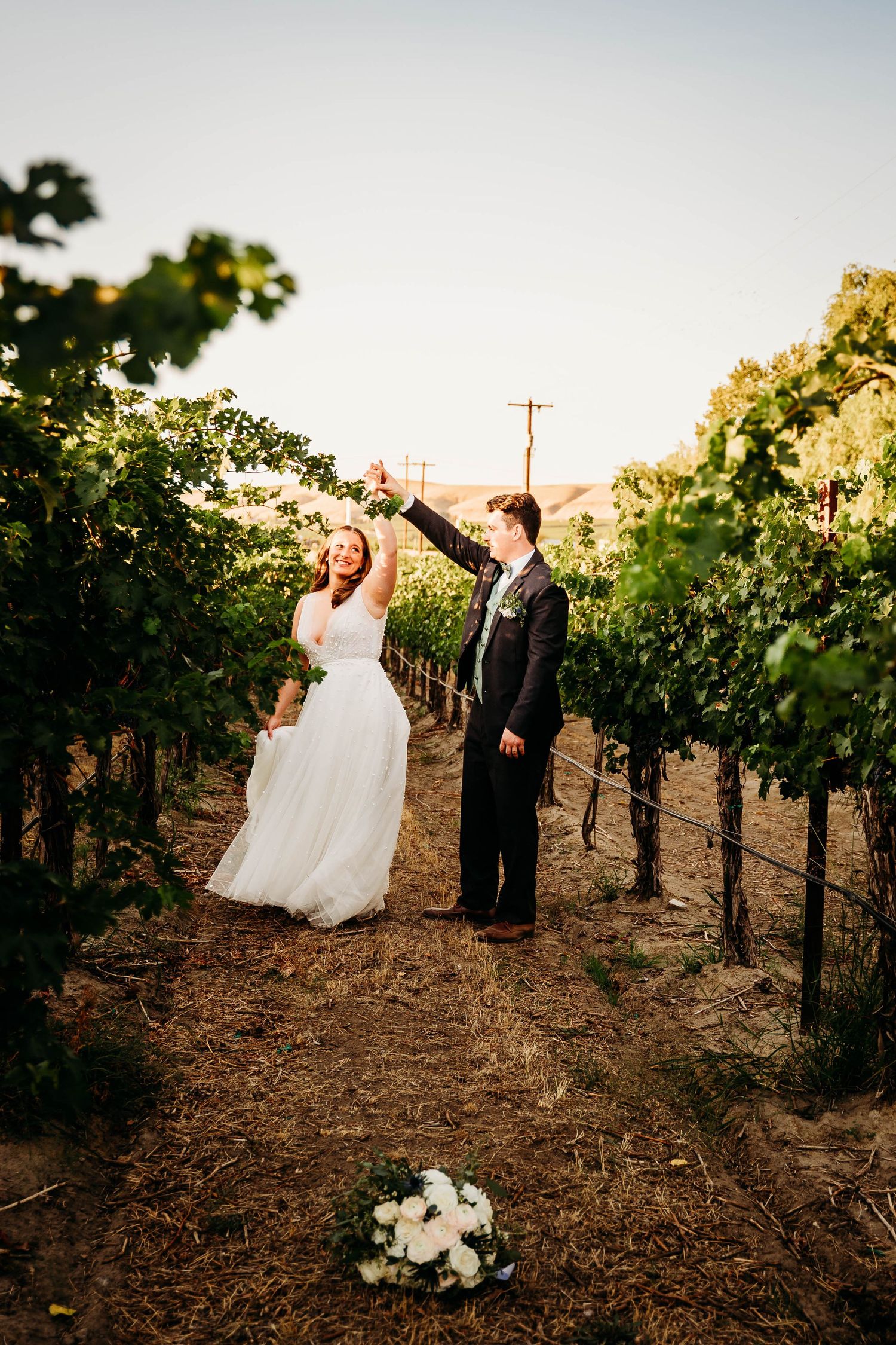 Joyful bride and groom raise hands in celebration while dancing in a vineyard.