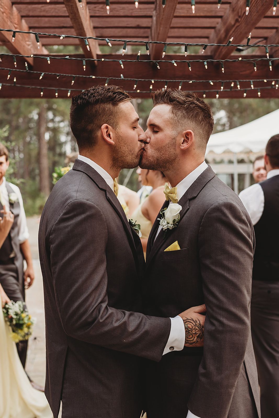Two grooms sharing a first kiss at their outdoor wedding ceremony under string lights.