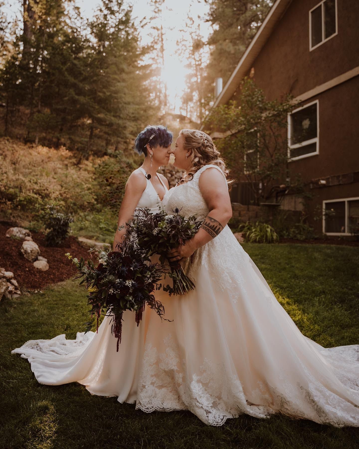 Two brides in white lace gowns hold dark floral bouquets and touch foreheads in golden sunset backyard light.