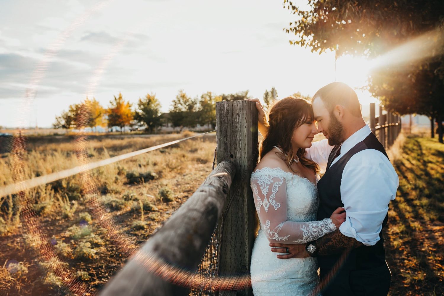 Couple embracing near wooden fence at golden sunset in a rustic outdoor setting.
