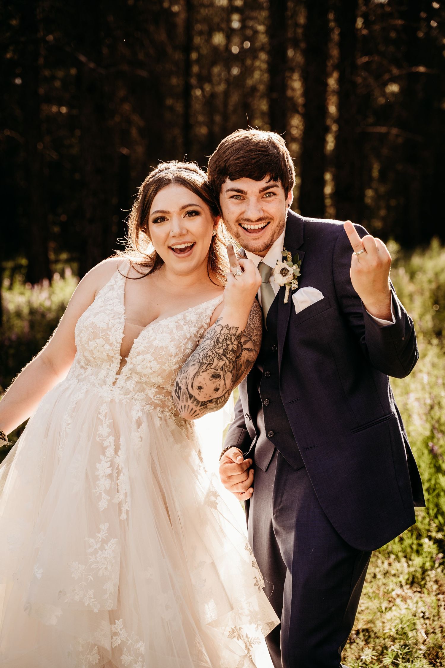 Ecstatic bride and groom laugh together showing wedding rings, bride with tattoo in lace gown in sunny forest.
