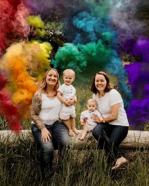 Two women with babies posing with colorful rainbow smoke bombs outdoors.