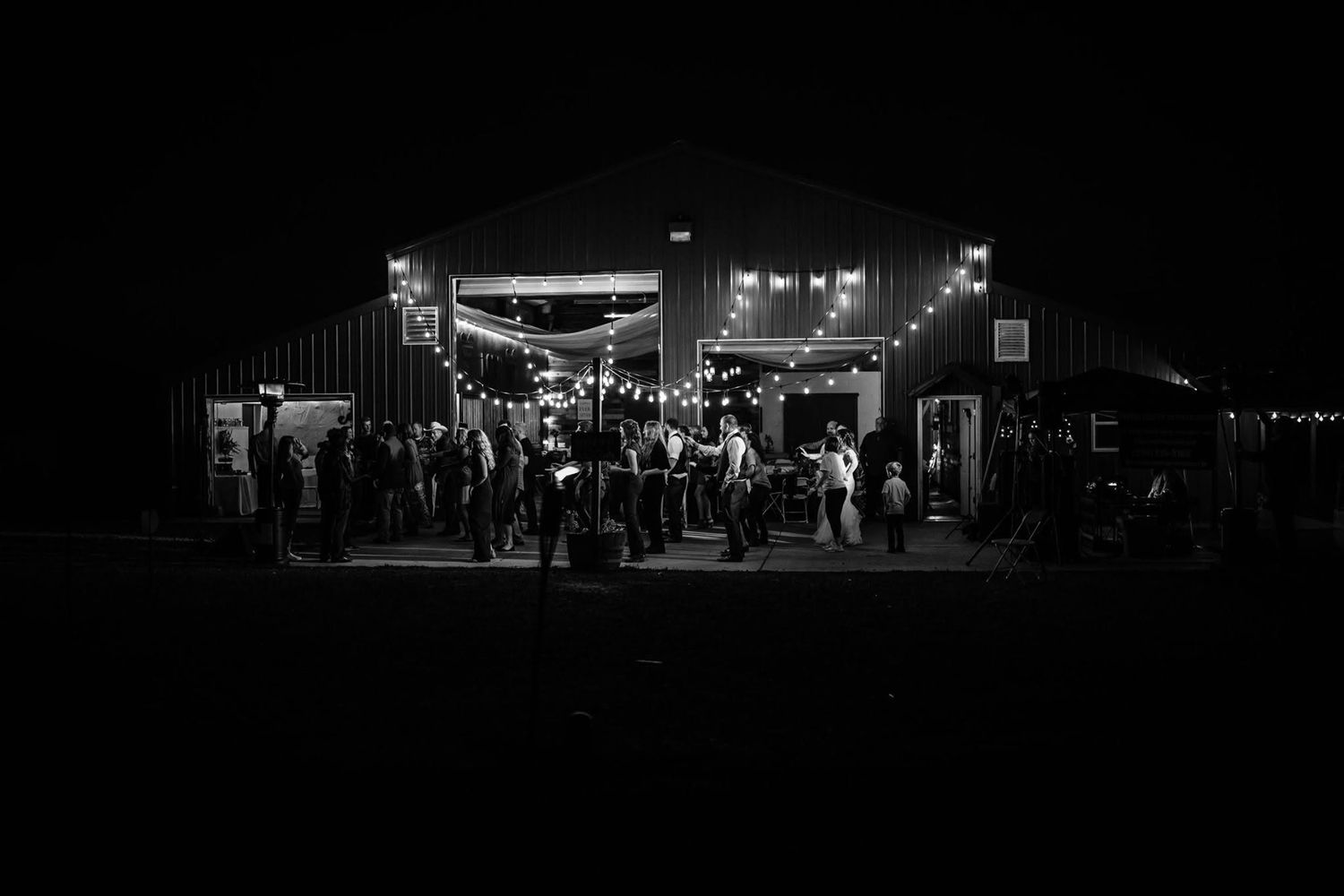 Black and white photo of wedding guests gathering outside a barn venue at night.