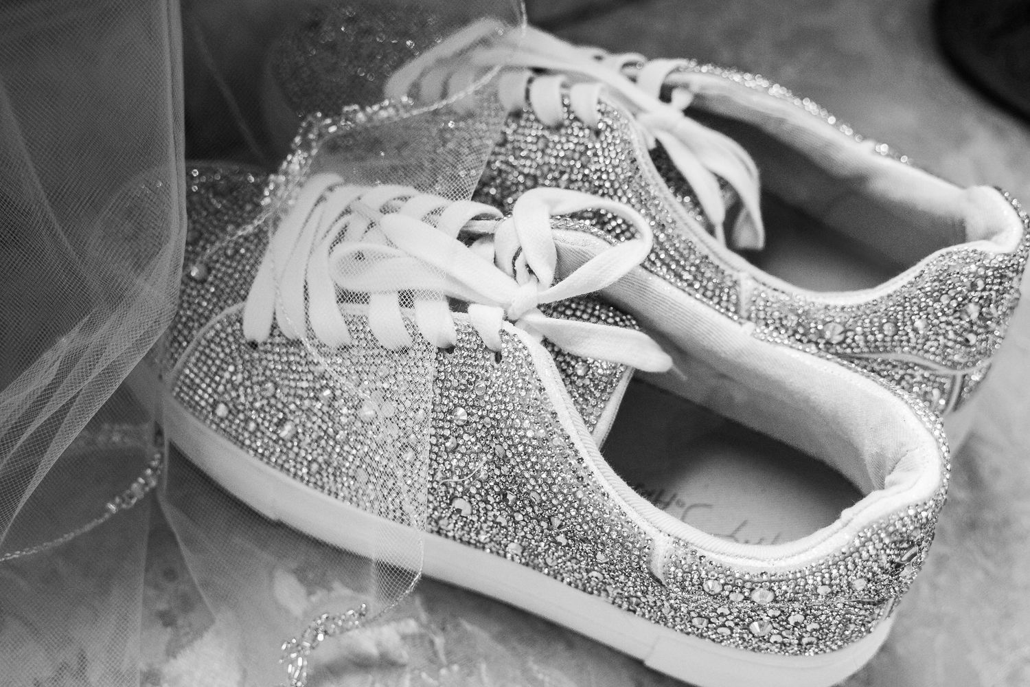 Black and white close-up of sparkling crystal rhinestone bridal sneakers beside flowing wedding veil.